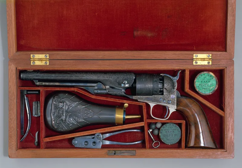 Wooden case lined in red velvet holding an engraved Colt revolver with a walnut grip, a powder flask, a bullet mould, round tins of caps and small tools.