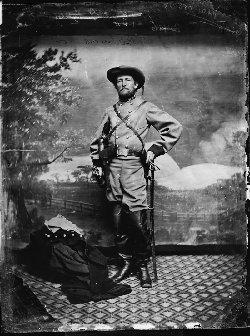 Full-length photograph of Mosby, bearded, in a plumed slouch hat and a grey shell jacket with cross-belts, one hand on his hip and the other on a sword, before a painted landscape backdrop.