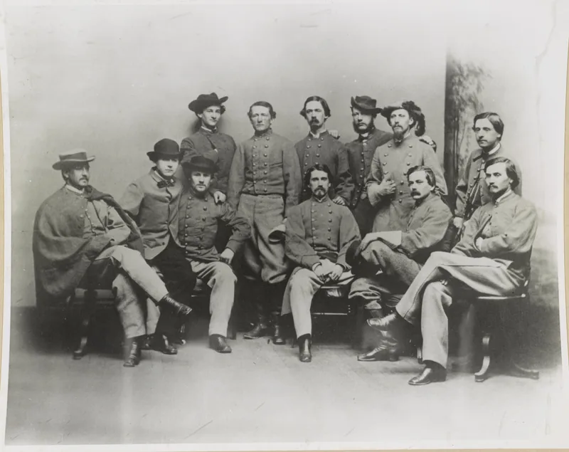 Studio group portrait of eleven men in grey double-breasted jackets and slouch hats, some seated with crossed legs, one wrapped in a cape, against a painted backdrop.