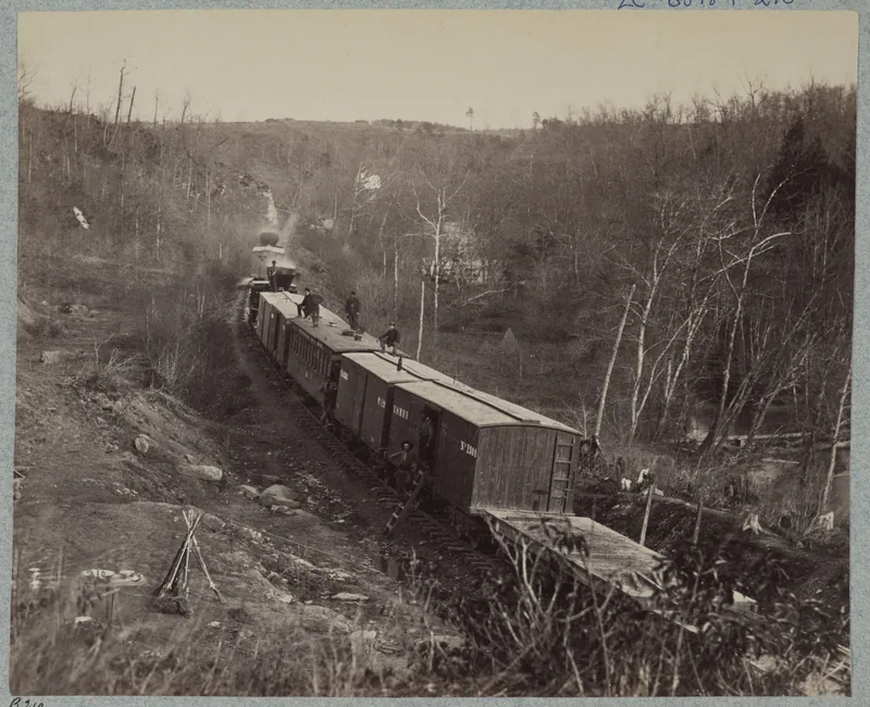 Photograph of a train of wooden boxcars curving through a cutting in leafless woods, men sitting on the car roofs, with rifles stacked in a tripod in the foreground.